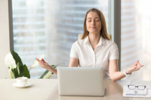Young woman meditating while at work with a laptop in front of her and a coffee cup on the table.