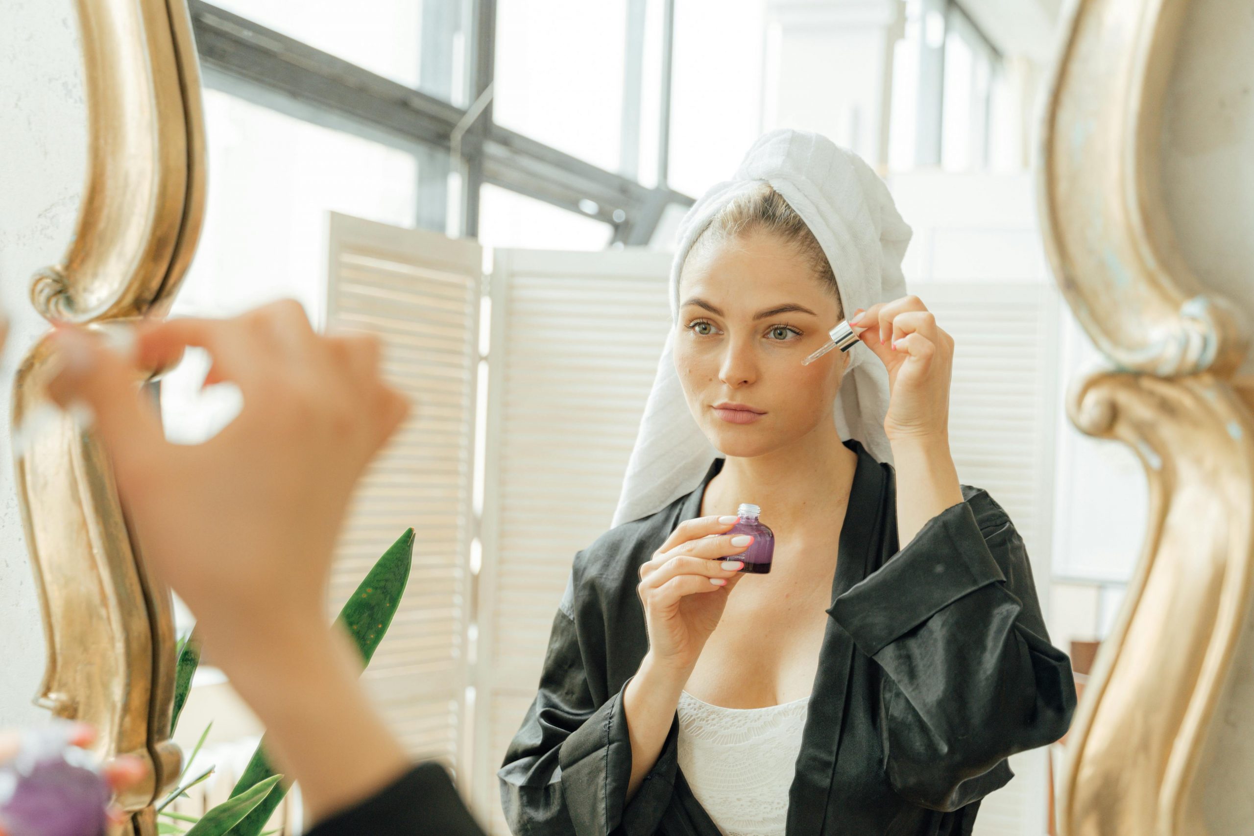 woman with a towel wrapped around her head applying retinol serum with a dropper in front of a mirror